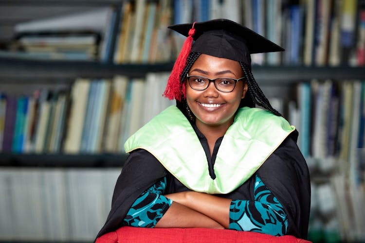 Smiling Woman In Glasses In Graduation Gown Sitting With Bookcase In Background