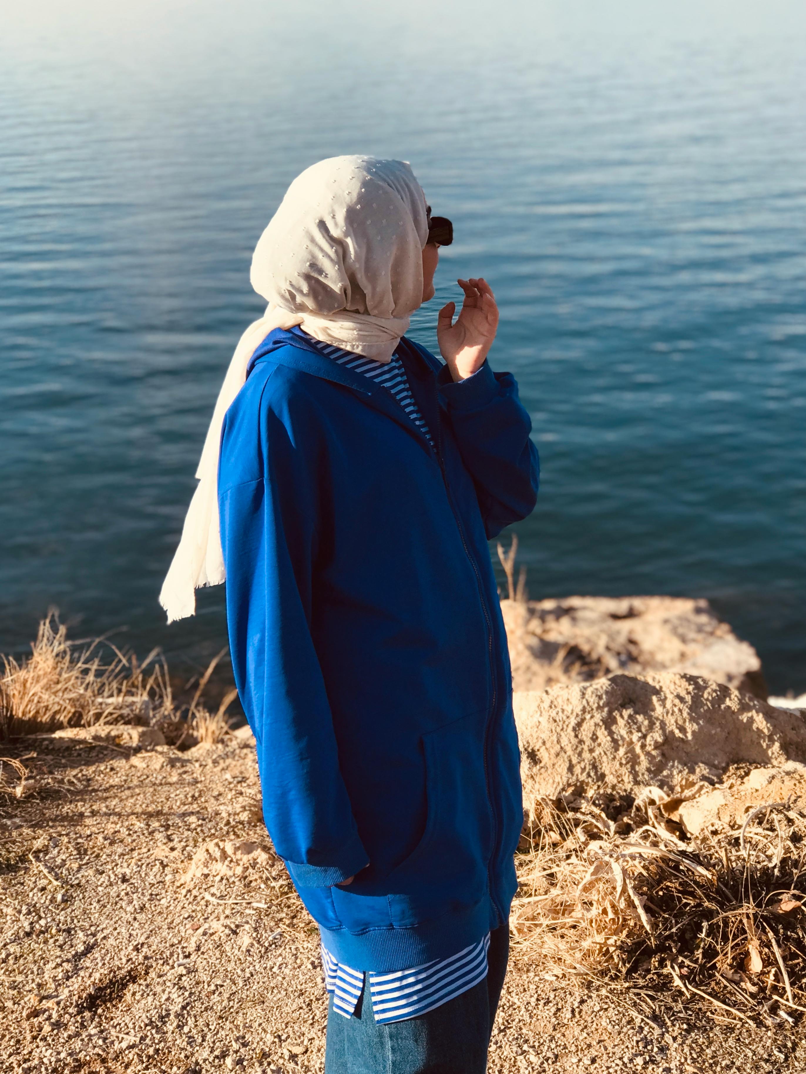 A woman wearing a hijab and blue jacket gazes at a tranquil lake, creating a peaceful scene.