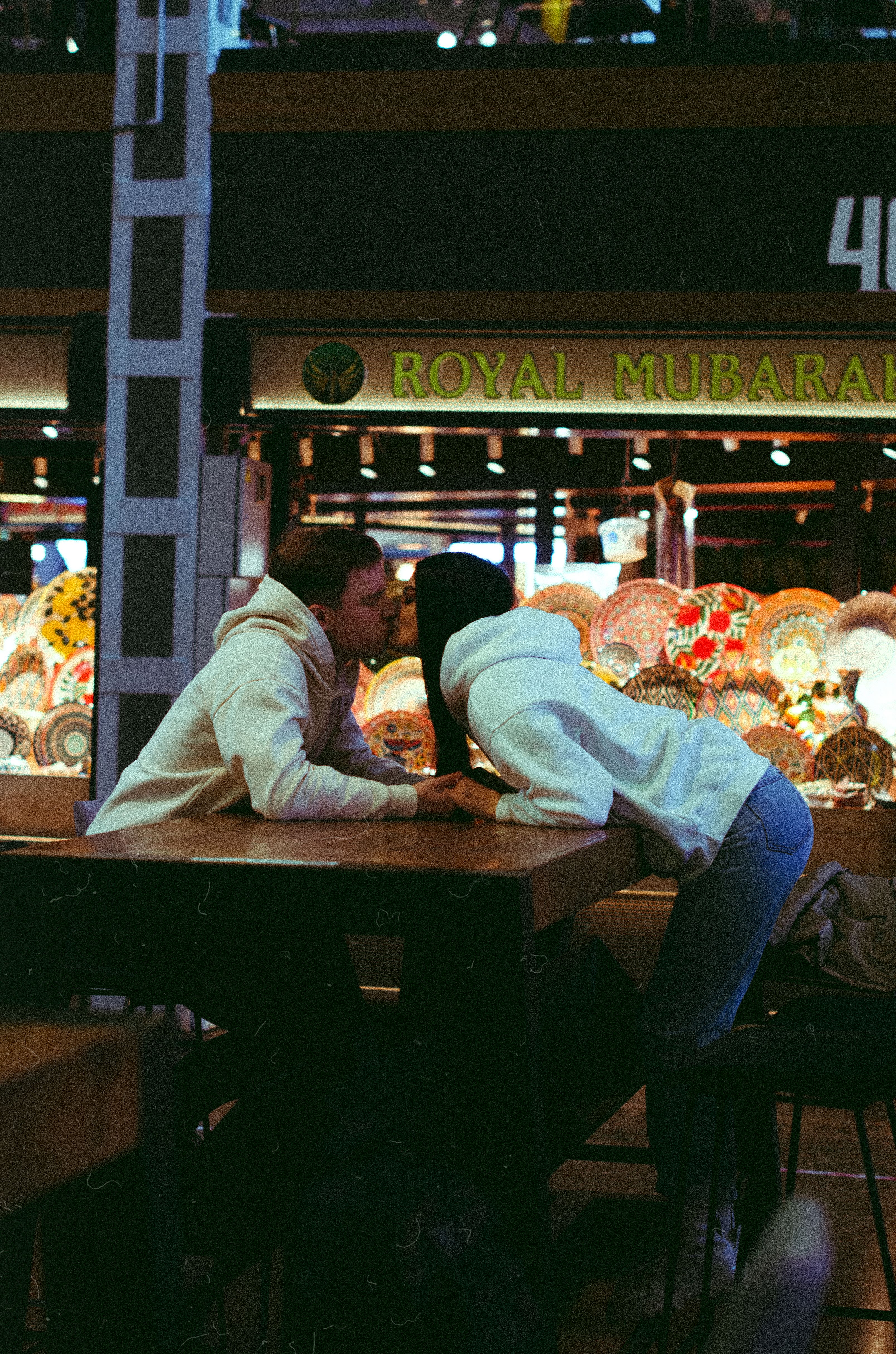 A couple shares a kiss at a marketplace table, surrounded by colorful lamps.