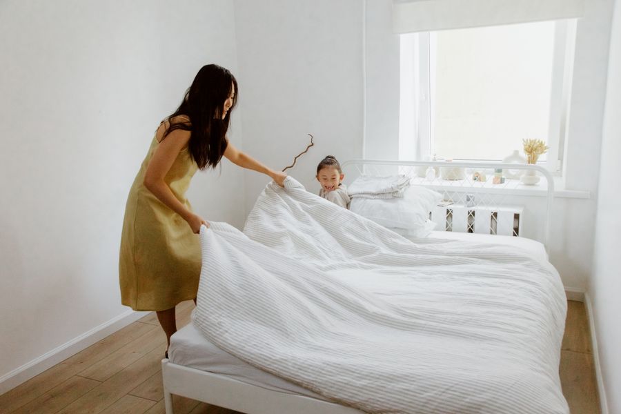 A mother and daughter making the bed in a bright, airy bedroom with white walls.