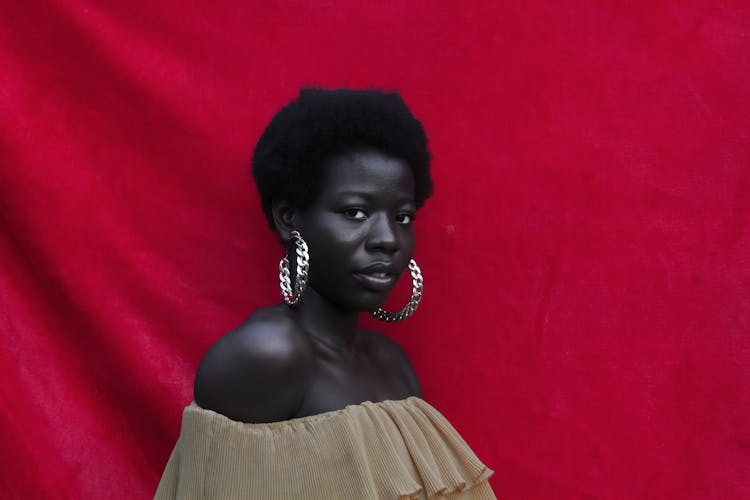 A Woman In A Beige Off Shoulder Top Beside A Red Curtain