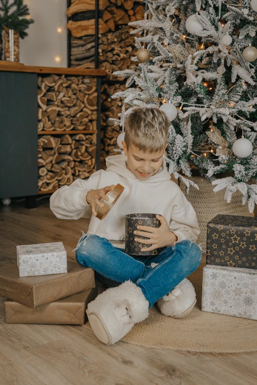 Boy in White Hoodie Opening Gift Beside Christmas Tree 