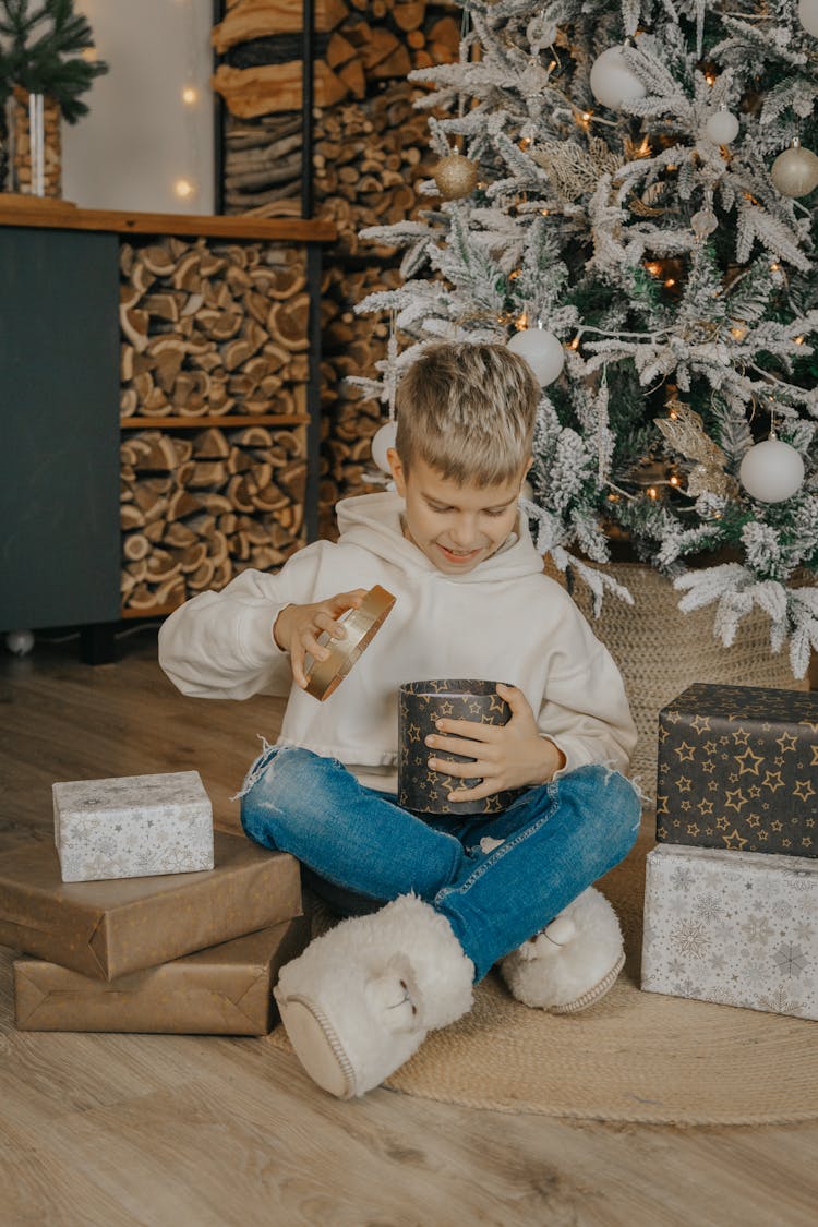 Boy In White Hoodie Opening Gift Beside Christmas Tree 