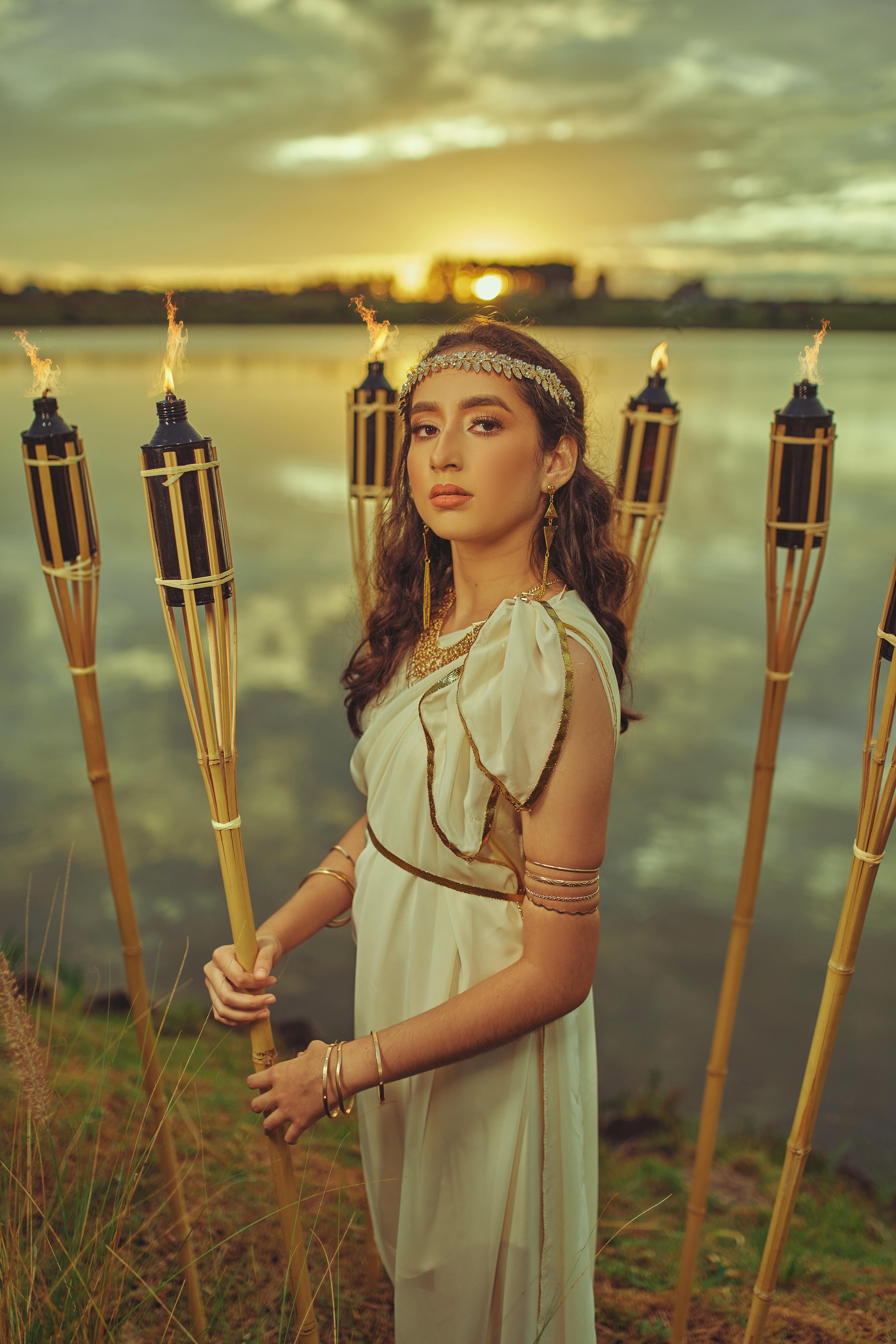 A woman in a white dress poses by a lakeside with torches at sunset, evoking a serene and mystical atmosphere.