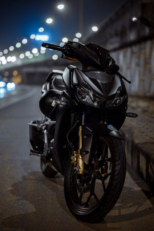Free A modern black motorcycle parked on a dimly lit urban road, showcasing its sleek design and headlight details. Stock Photo