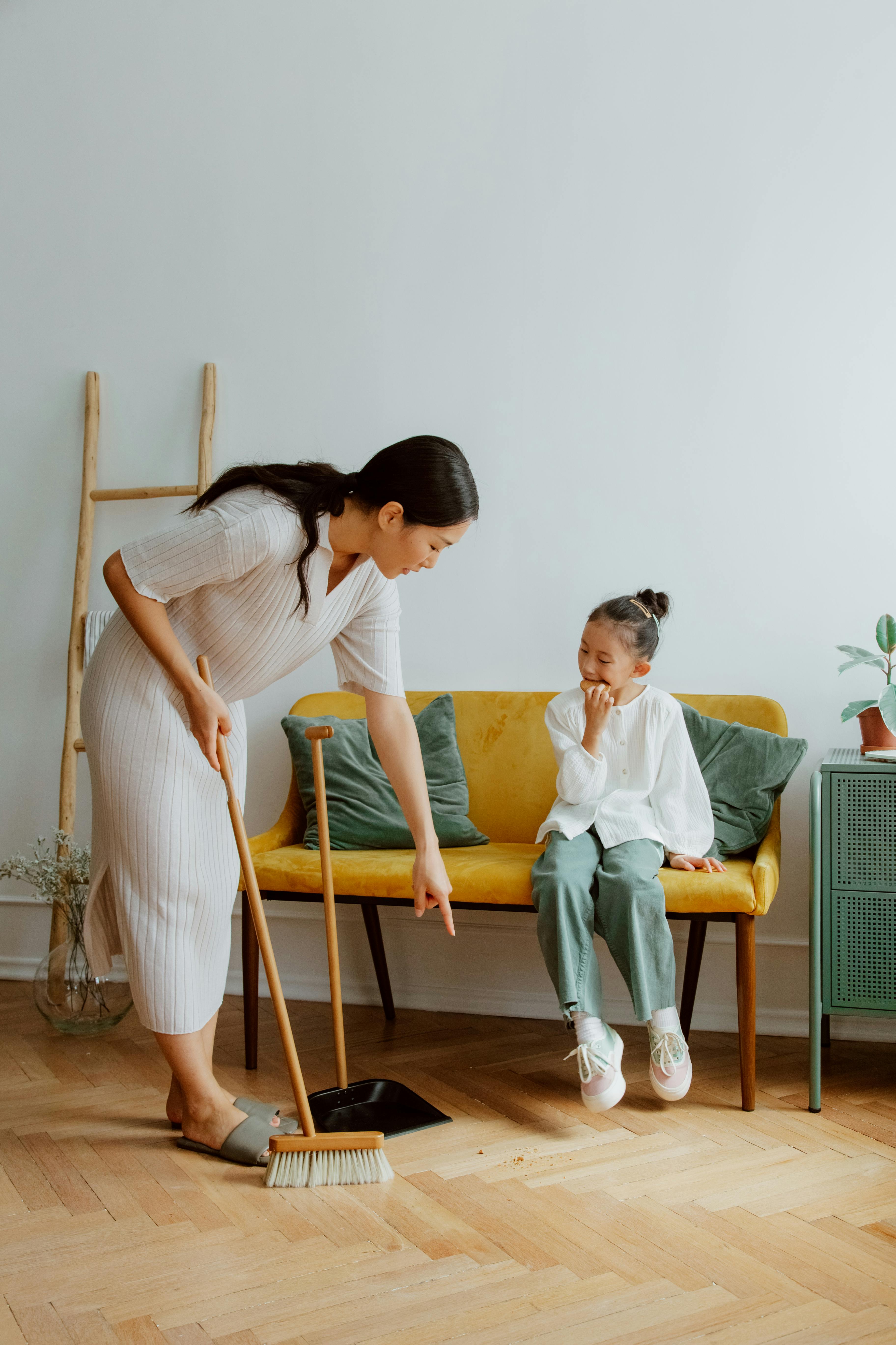 mother sweeping floor and daughter sitting on sofa