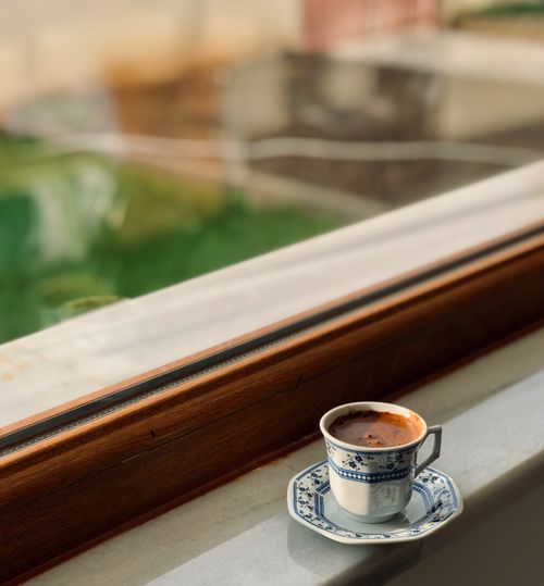 Free A intricate coffee cup sits on a windowsill with a sunny outdoor view. Stock Photo