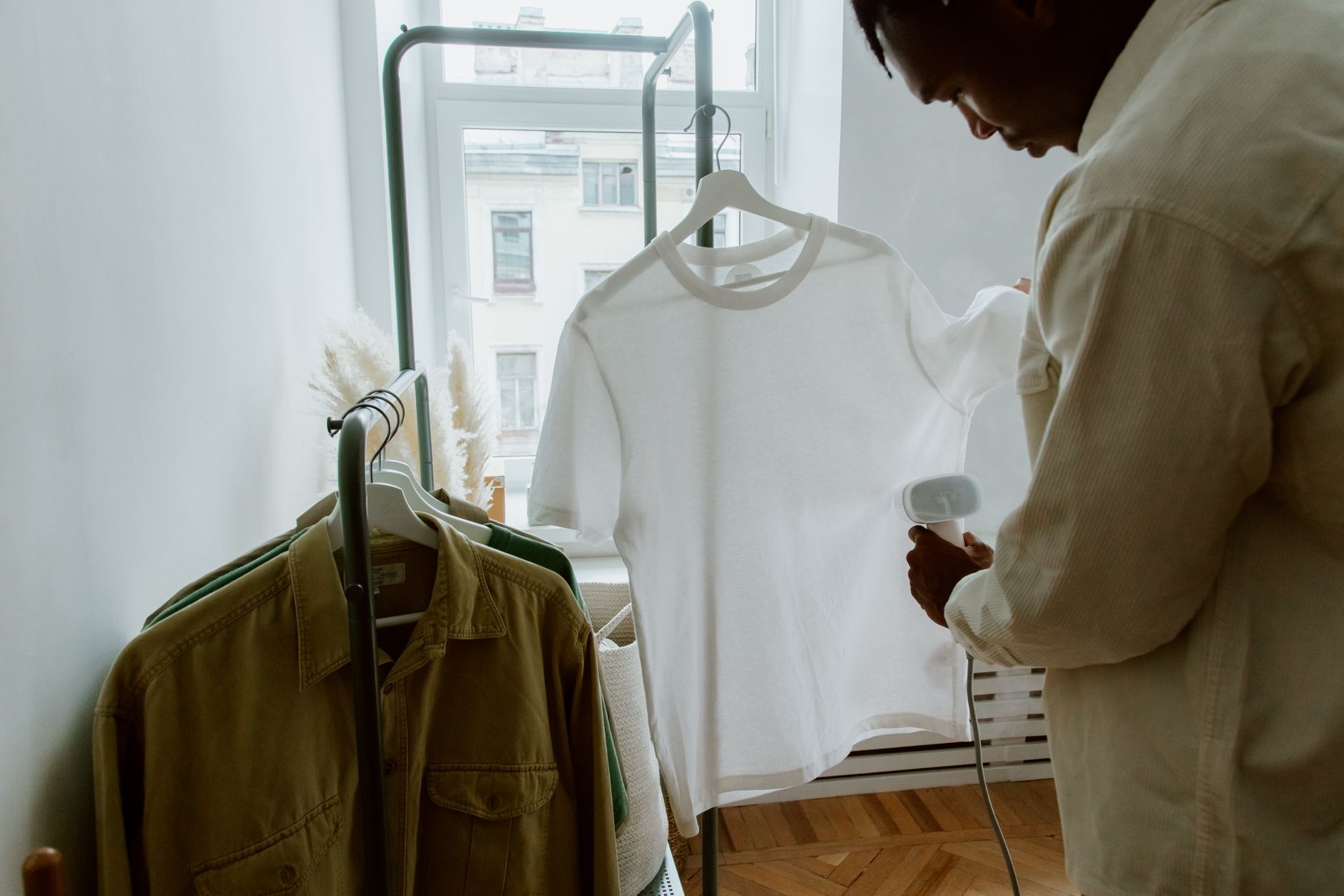 How To Iron A Dress Shirt Without Shine Marks 4 A man uses a steam iron on a white shirt hanging from a clothing rack indoors.
