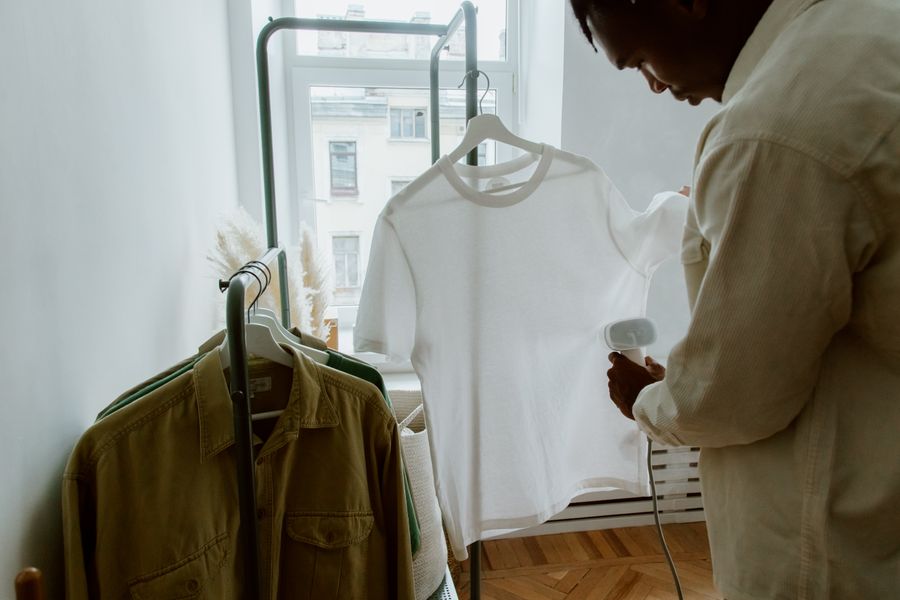 A man uses a steam iron on a white shirt hanging from a clothing rack indoors.