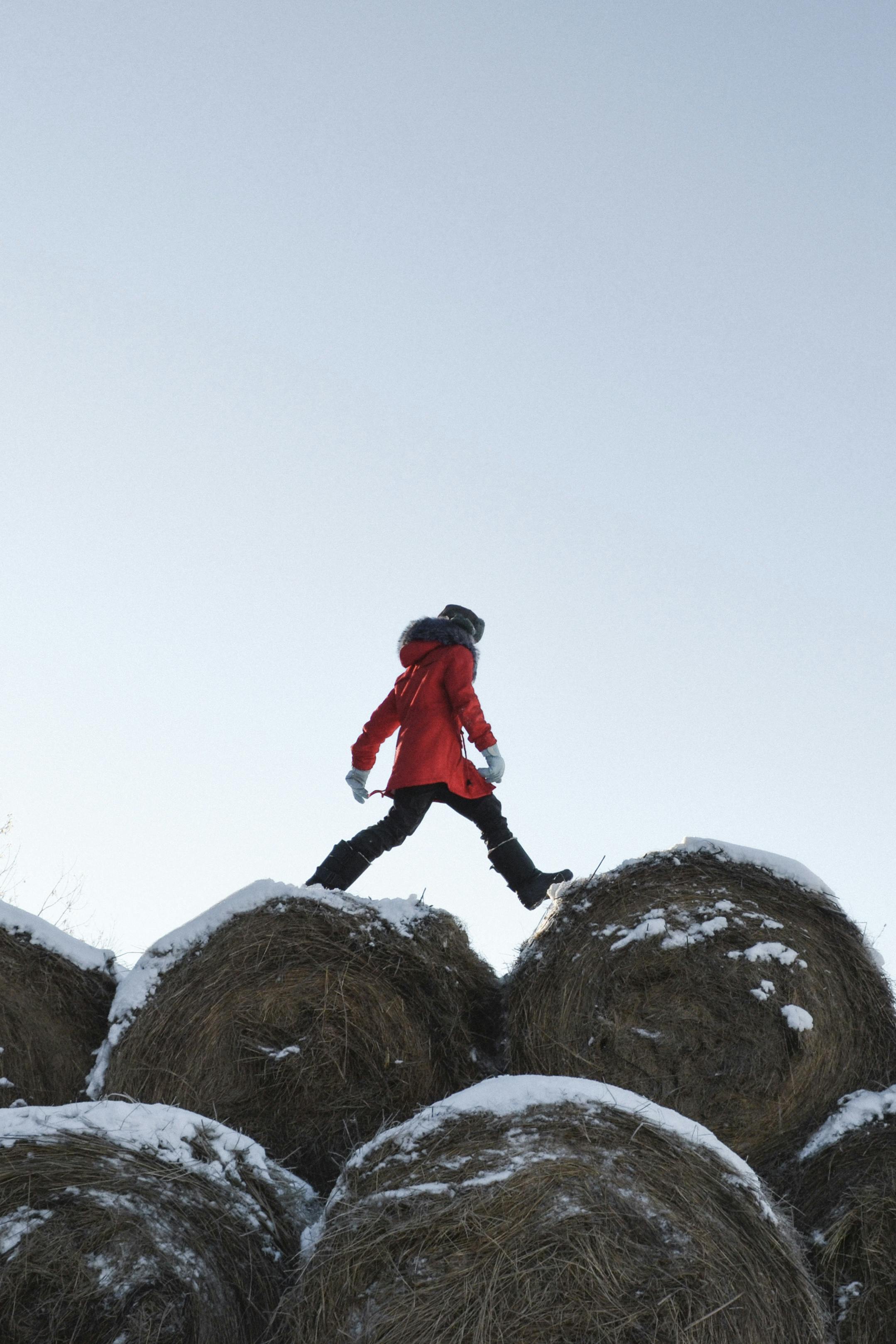 A person in a red jacket walks on snowy hay bales against a clear sky.
