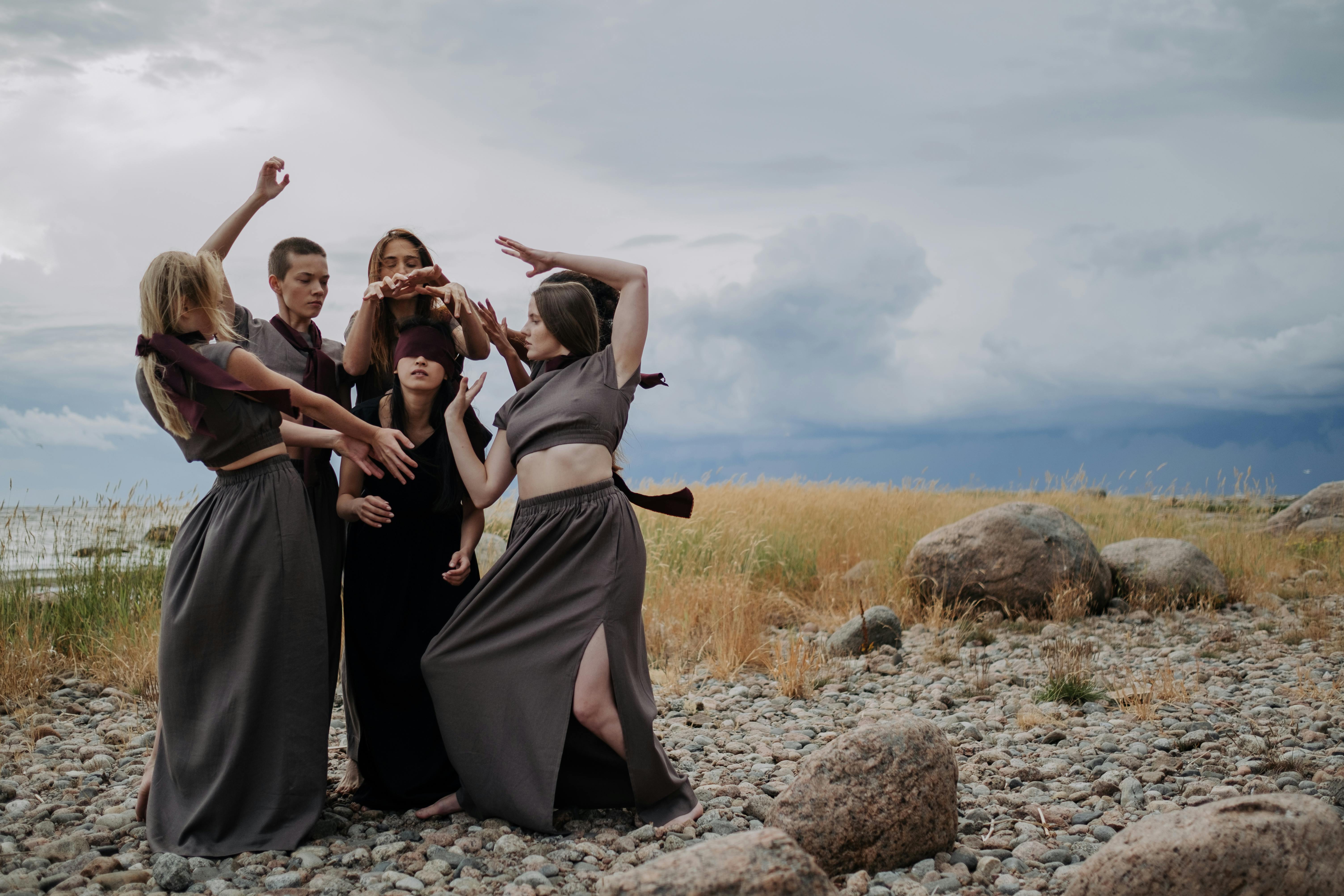 a blindfolded person surrounded by women wearing long dresses