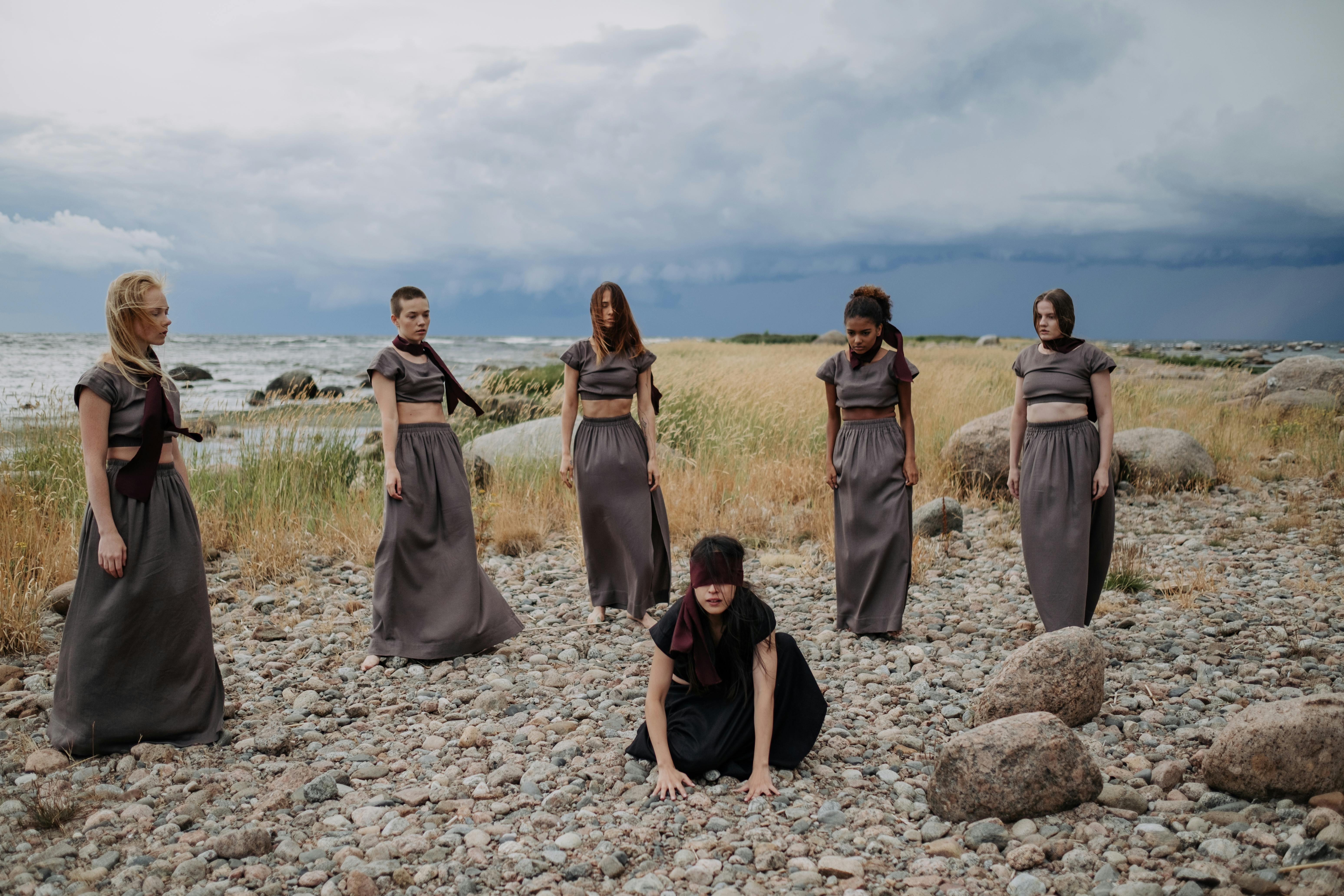 blindfolded woman kneeling on the ground surrounded by a group of women in gray dresses