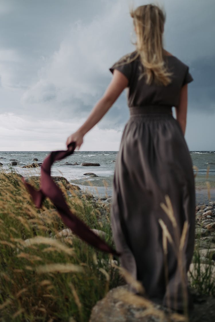 A Woman In Gray Long Dress Standing On Big Rock While Holding A Fabric