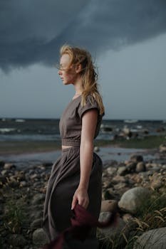 A woman stands by a rocky seashore under a moody, stormy sky, embodying solitude and introspection.
