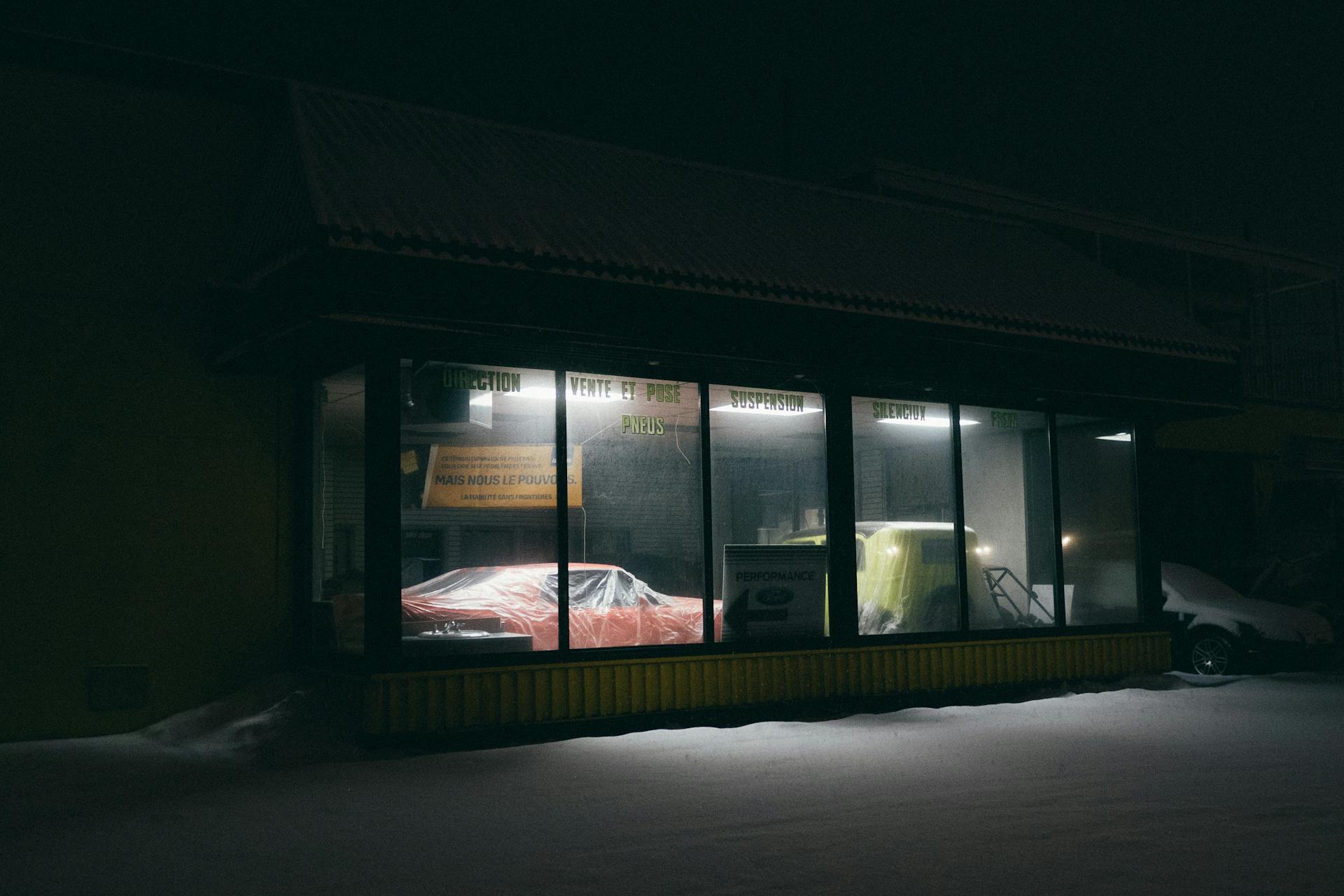 A night view of an auto repair shop with cars inside, seen through glass windows.