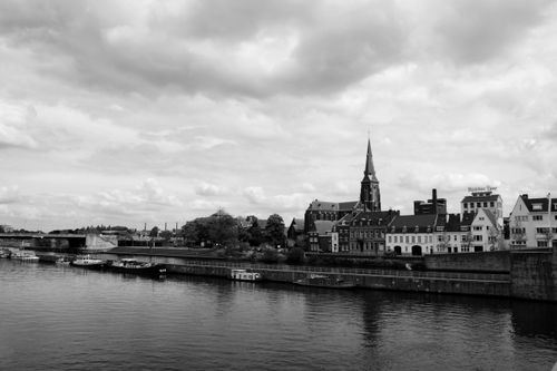 Free Black and white photo of Maastricht's riverfront showcasing historic architecture and a church steeple under a cloudy sky. Stock Photo