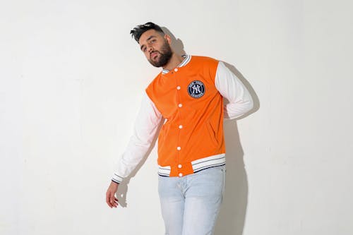 Man in Orange and White Zip Up Jacket and Blue Denim Jeans Standing Beside White Wall
