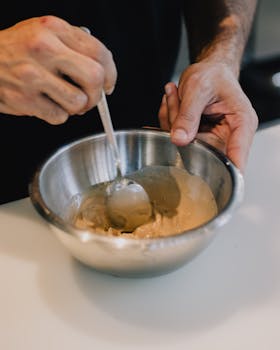 A person stirring creamy sauce in a stainless steel mixing bowl on a white kitchen surface.