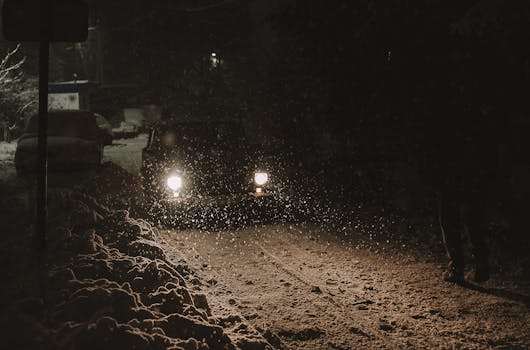 A car drives through a snow-covered road at night, with headlights illuminating falling snowflakes.