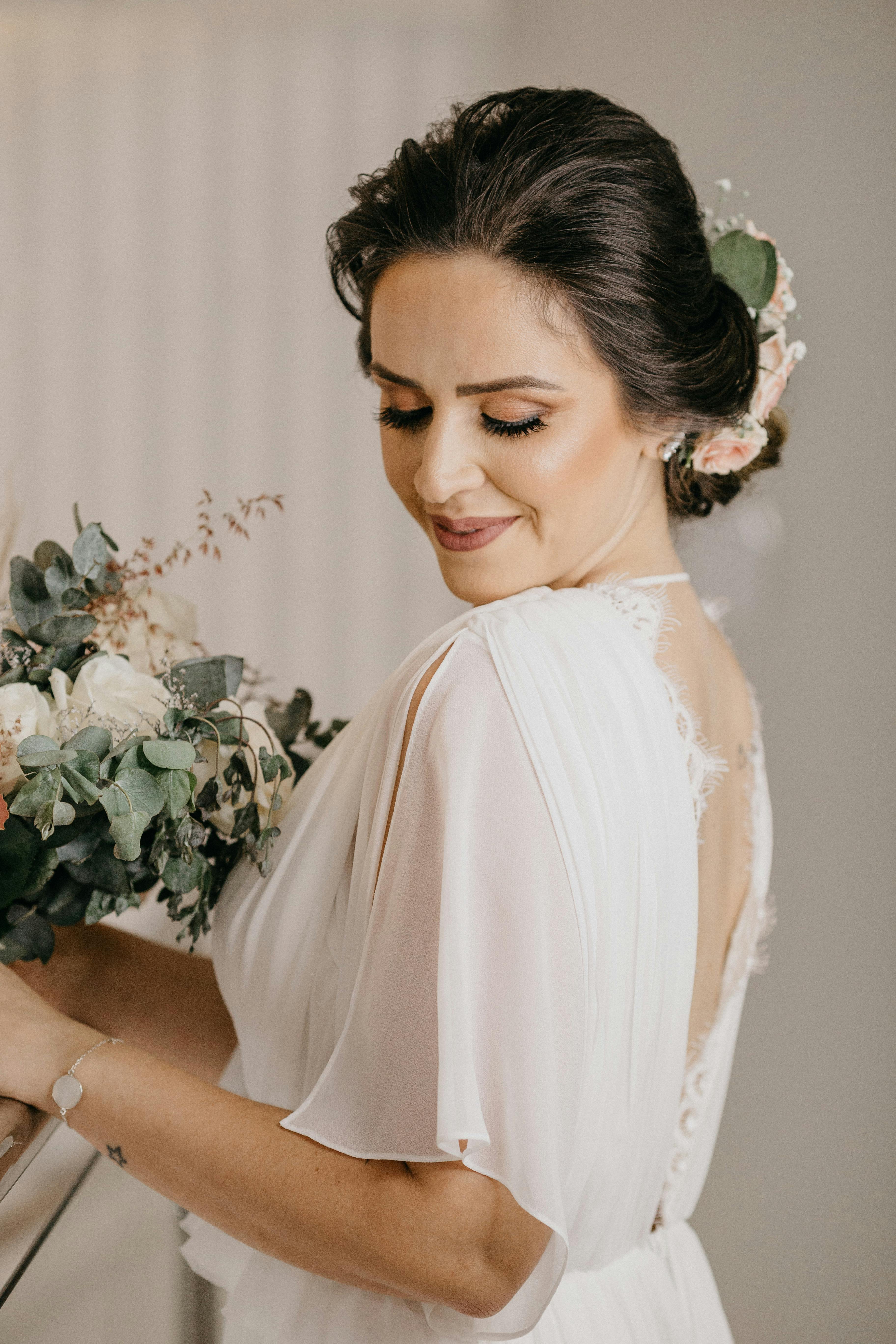 A beautiful bride in a white dress holding a bouquet, posing with a gentle smile indoors.