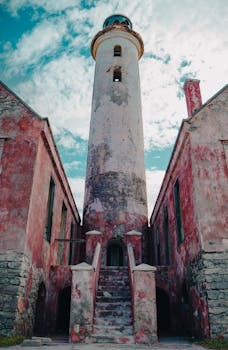 Vintage lighthouse with pink walls and weathered textures under a blue, cloudy sky.