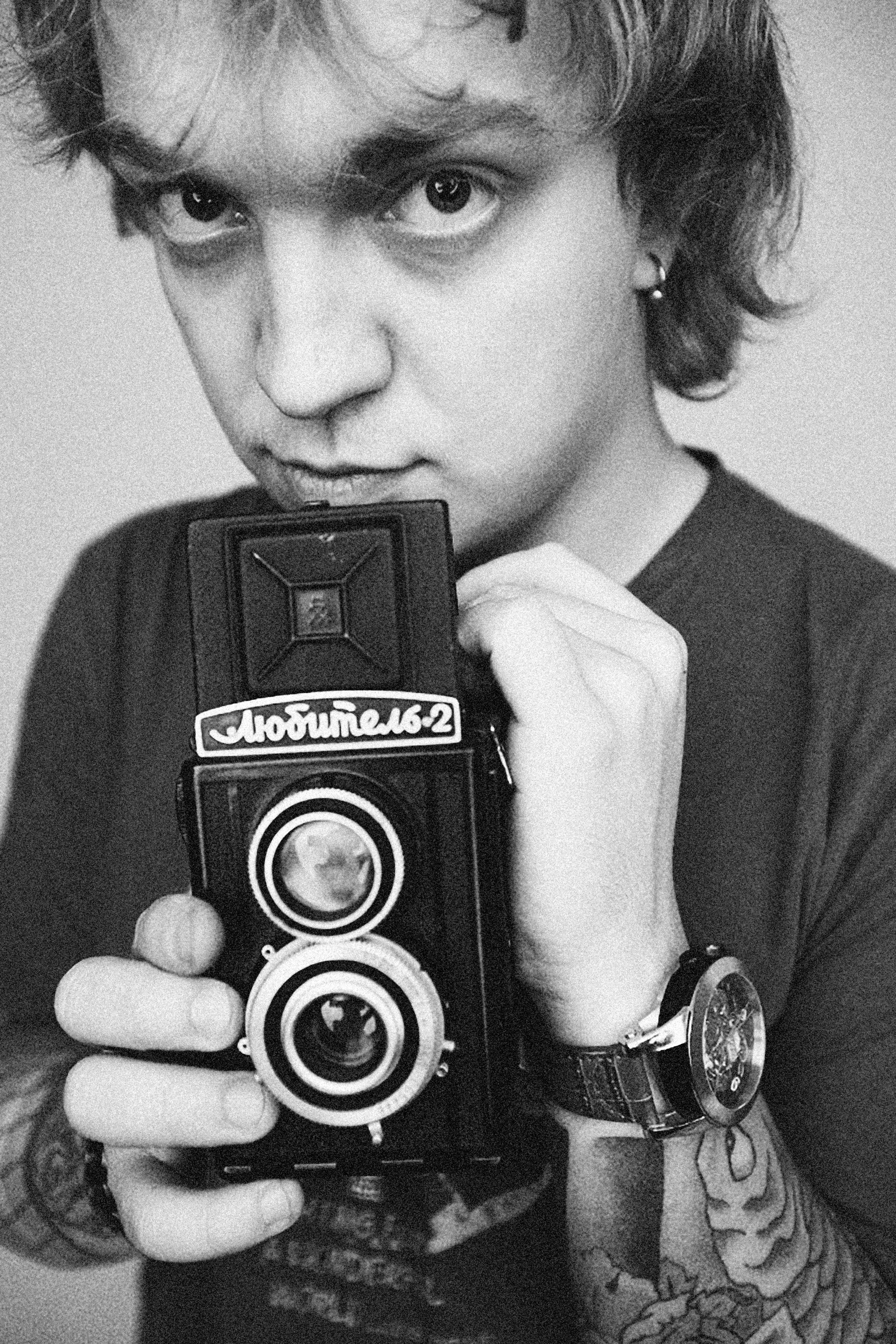 Black and white portrait of a tattooed adult holding an old twin-lens reflex camera, evoking nostalgia.