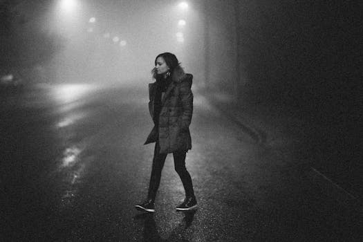 A woman walks alone on a foggy street at night, captured in a moody black and white style.
