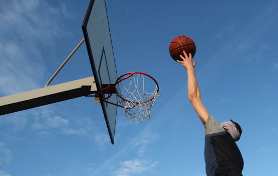 Dynamic action shot of a man jumping towards a basketball hoop under a clear blue sky.