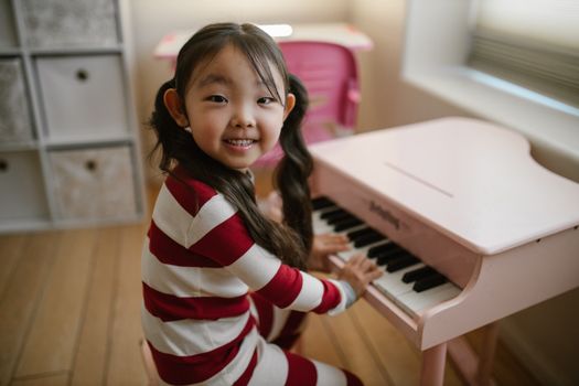 Smiling girl in pajamas playing a pink toy piano in a cozy home setting.