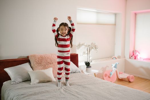 A joyful child in striped pajamas jumping on a bed in a bright and cozy bedroom setting.