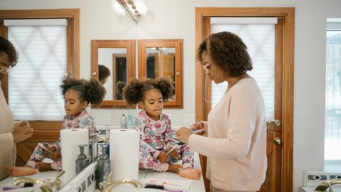 A mother assists her young daughter with her morning routine in a bright bathroom setting.