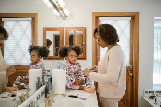 A mother assists her young daughter with her morning routine in a bright bathroom setting.