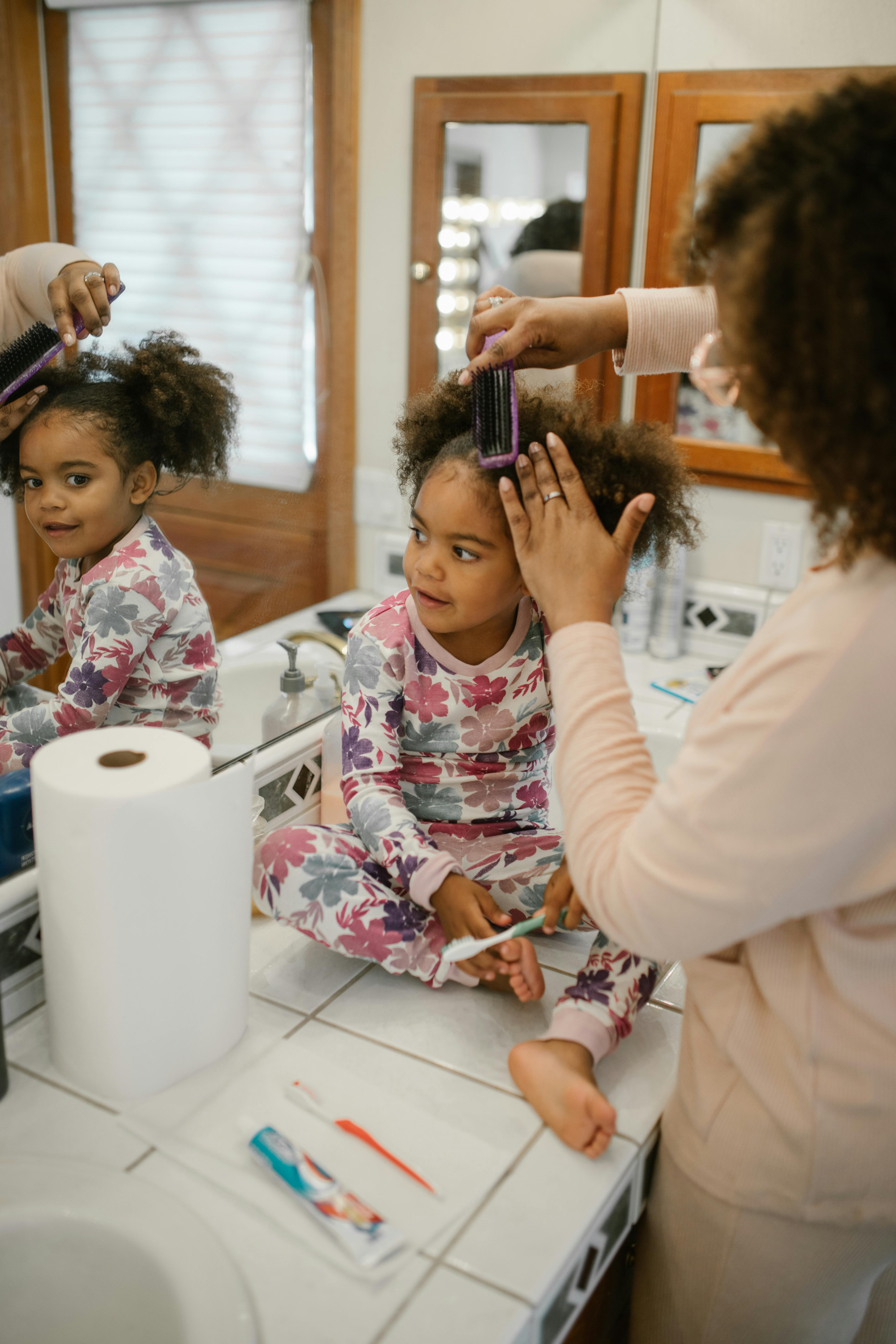 A mother styles her daughter's hair during their morning routine in the bathroom.