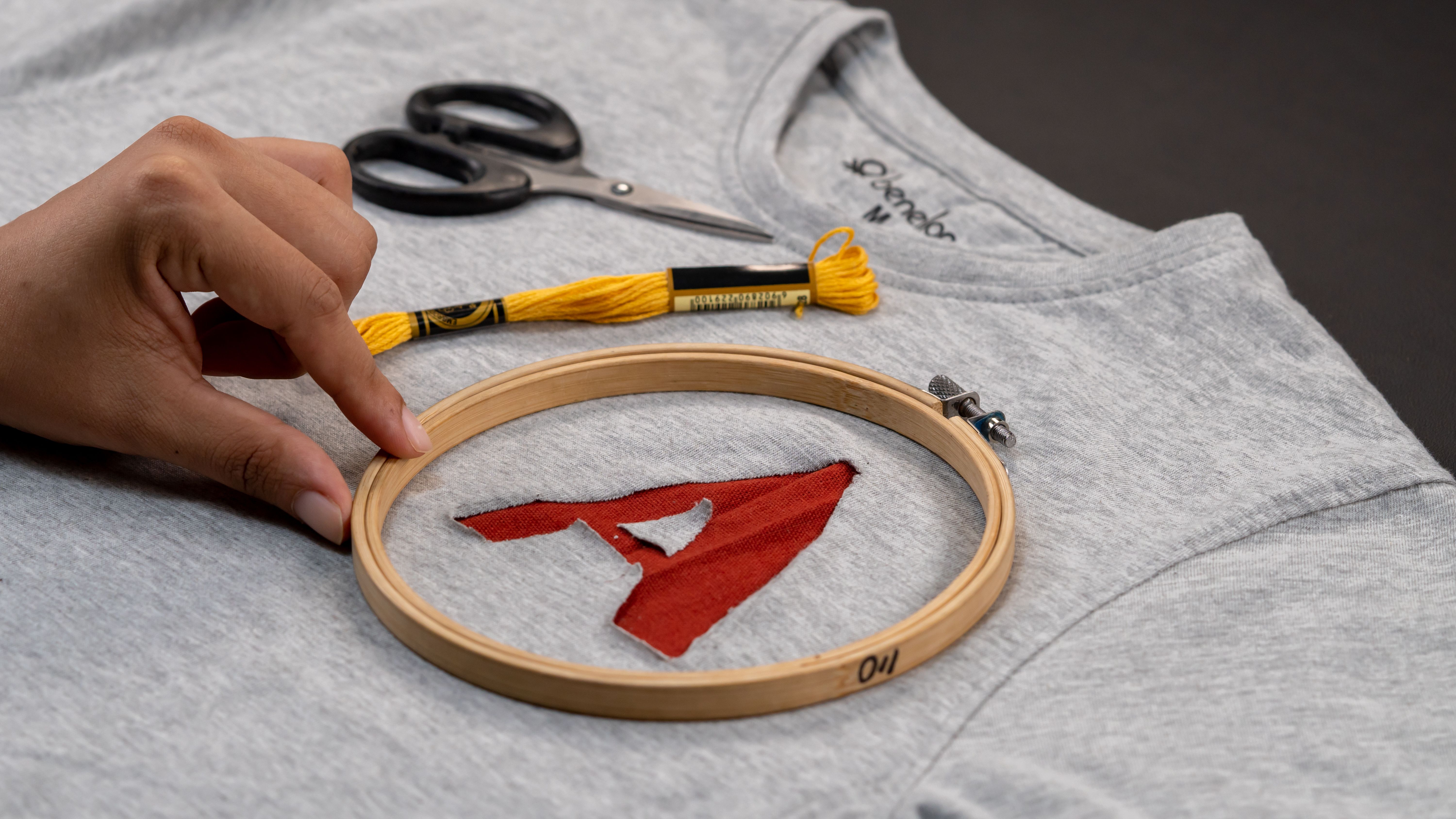 Close-up of hand embroidering a red letter on a gray sweatshirt using a wooden hoop.