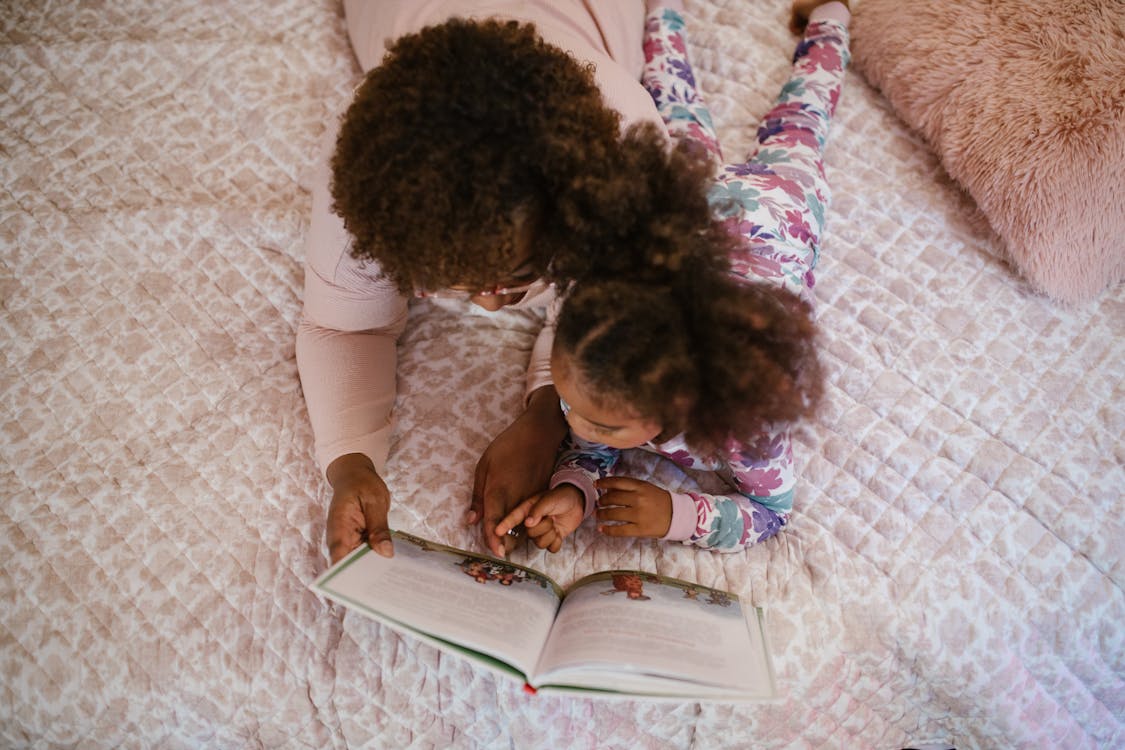 Mulher e criança leem um livro juntas, ambas estão deitadas na cama.