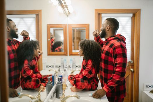 Father and daughter in matching pajamas brushing teeth together in a cozy bathroom.