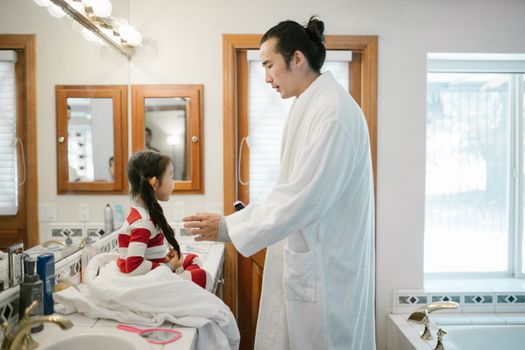 A father helps his daughter with her morning routine in a cozy home bathroom.