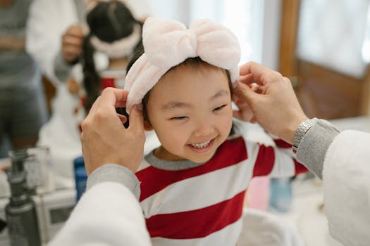 Child smiling while wearing a pink headband, being adjusted by an adult in front of a mirror.