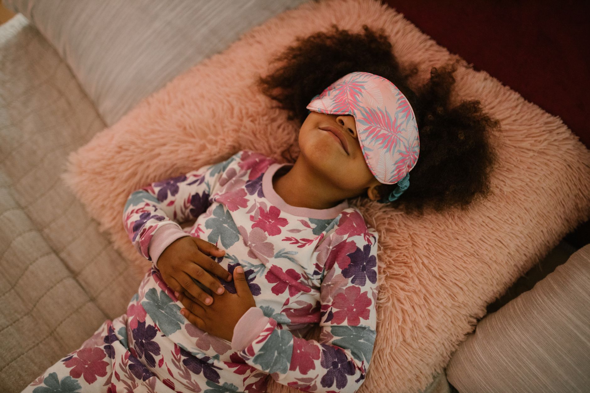 A young girl wearing floral pajamas and sleep mask, resting peacefully on a fluffy pillow.