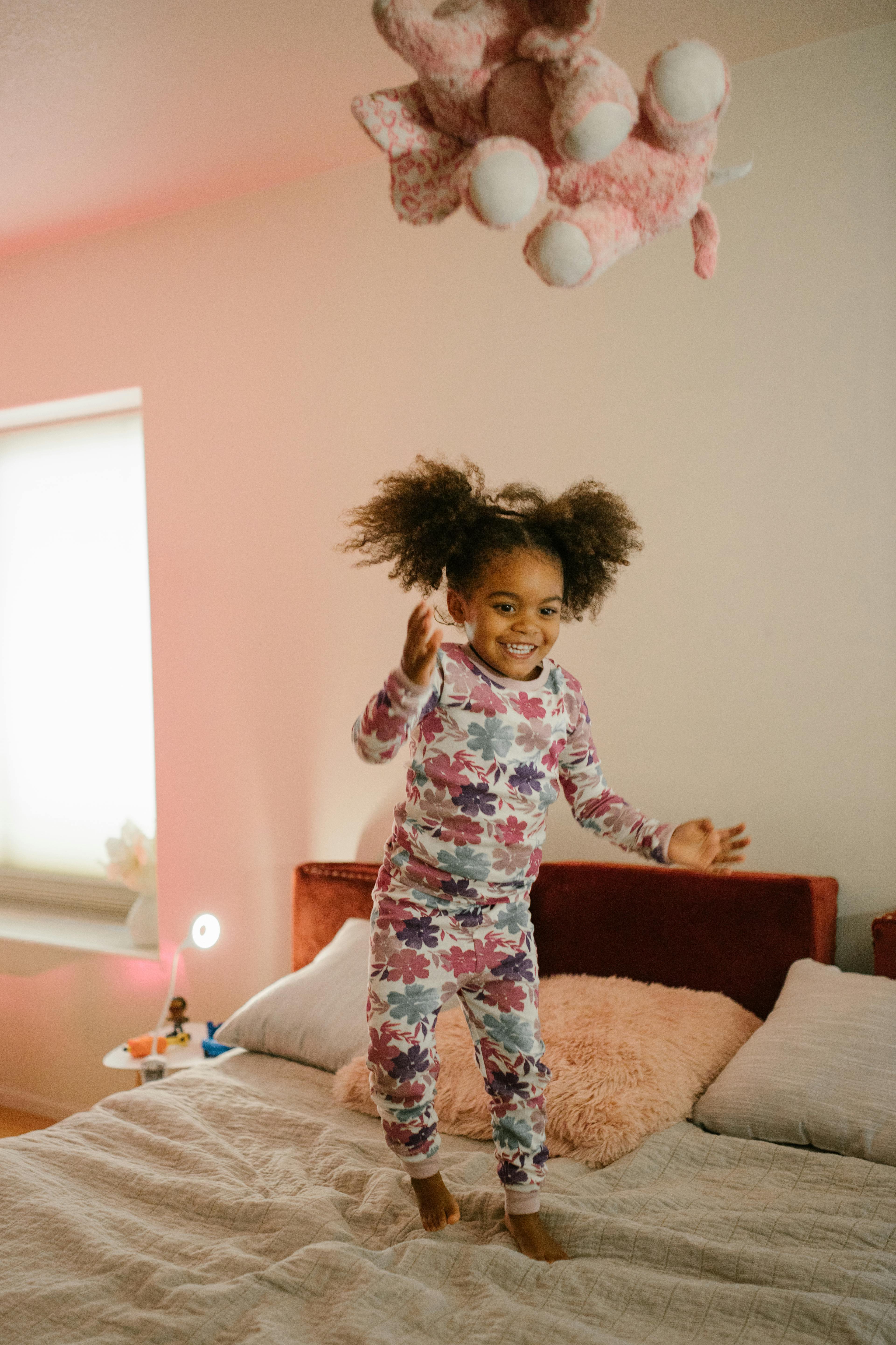 A happy child in pajamas jumping on a bed in a cozy, sunlit bedroom.