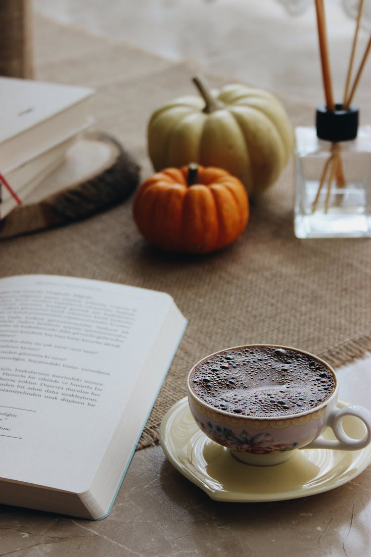 Cup Of Coffee Standing Next To Open Book And Pumpkins In Background