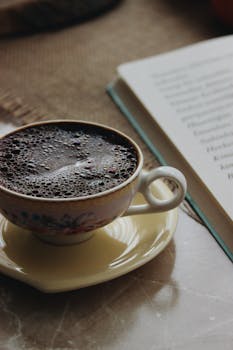 A warm cup of coffee on a saucer beside an open book on a table.