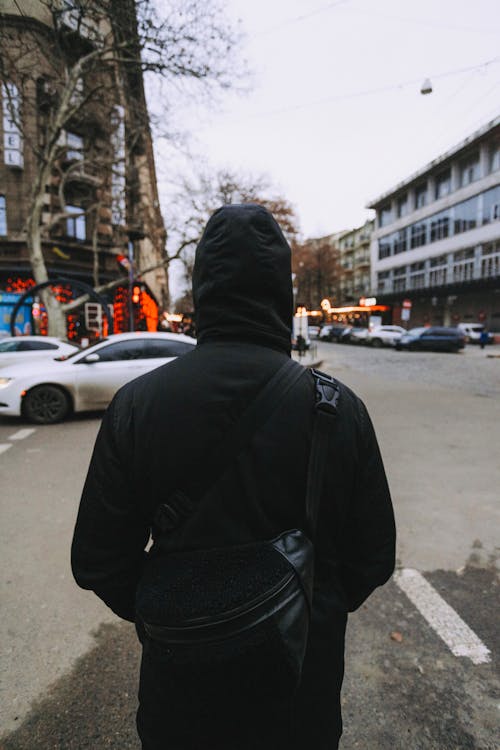 Free A Back View of a Person in Black Hoodie and a Cross Body Bag Standing on the Street Stock Photo