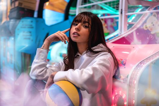 Stylish young woman sitting in a vibrant arcade holding a ball, surrounded by neon lights.