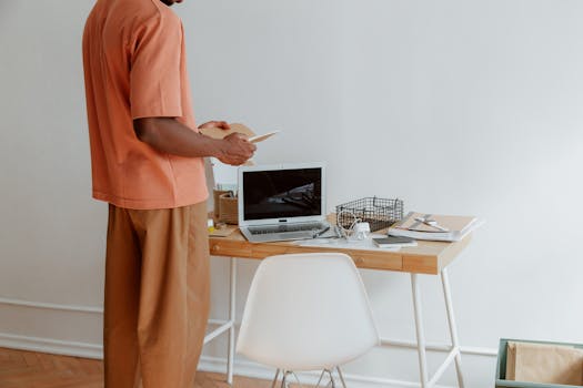 A person standing at a wooden desk with a laptop and office supplies. Minimalist aesthetic.