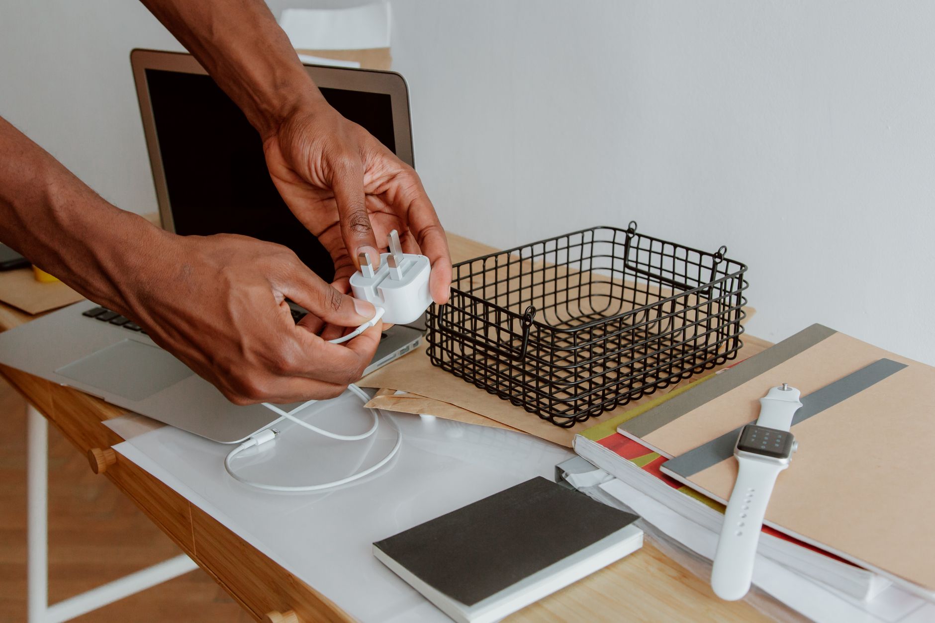 手機桌面佈置升級:App收納、Widget與走線全指南 Close-up of a man organizing a desk, handling a charger next to a laptop, wire basket, and notebooks.