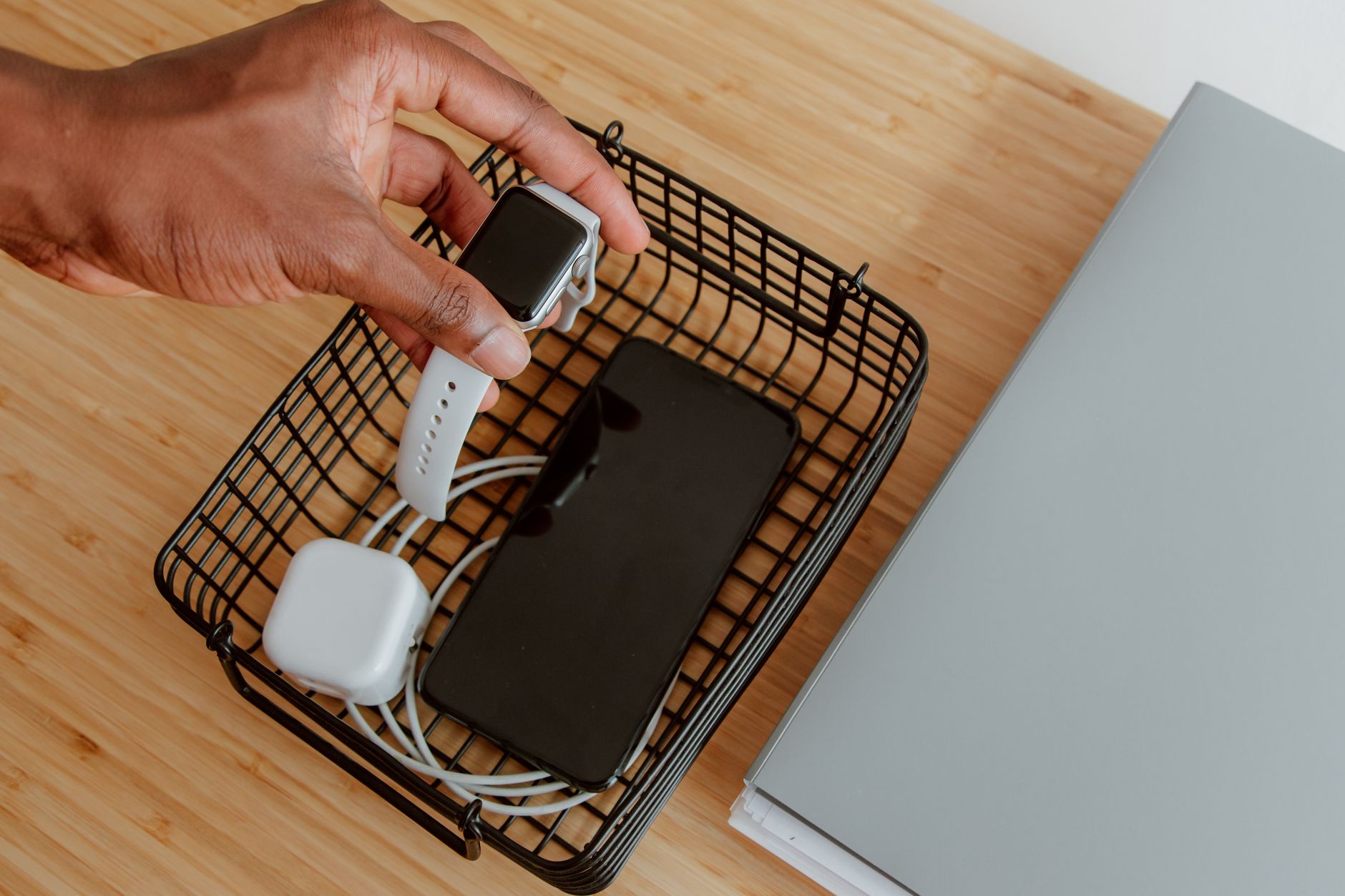 A hand lifting a smartwatch from a charging basket, with a smartphone and notebook nearby on a desk.