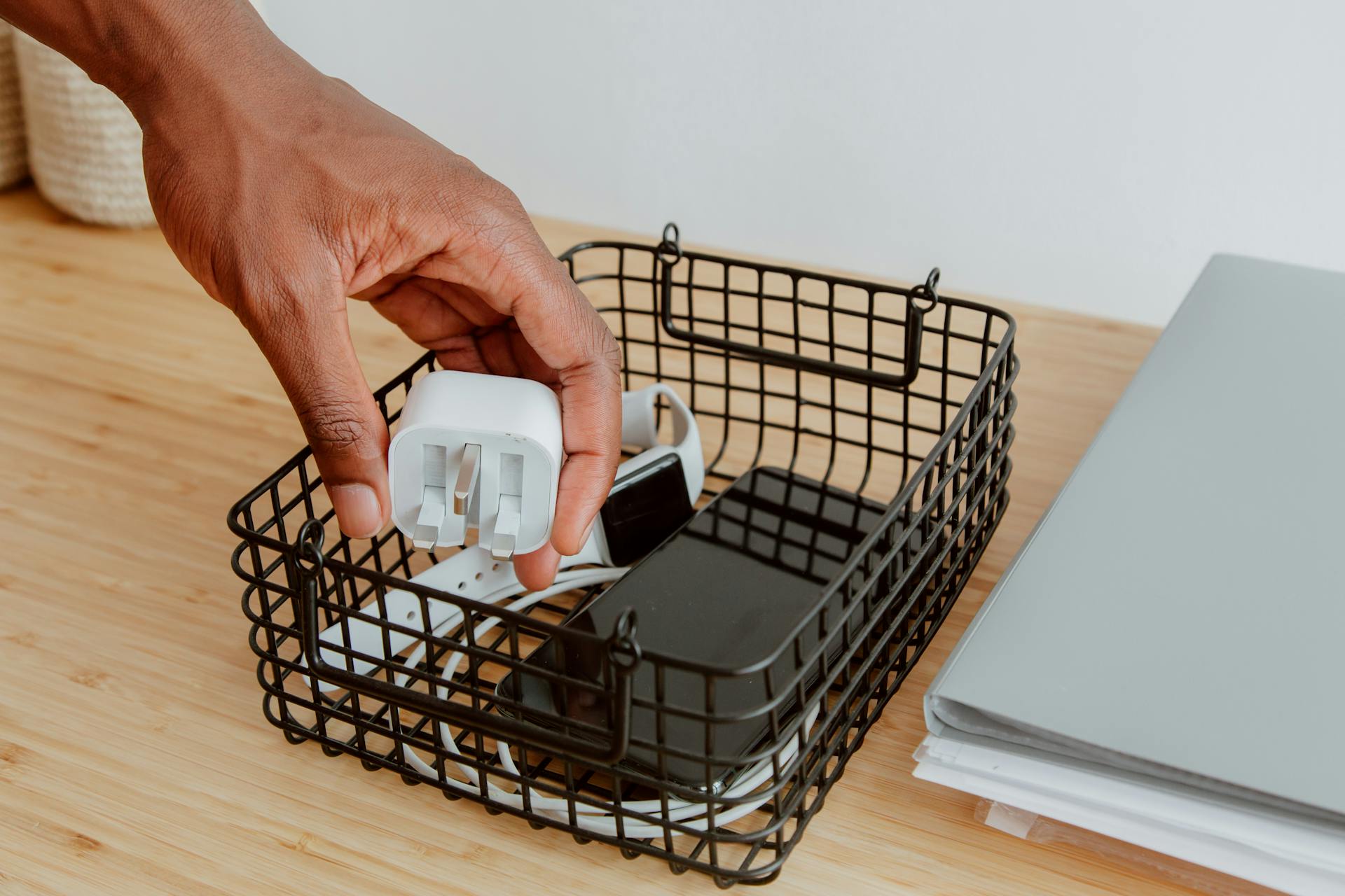 A hand organizing a desk with a basket of electronics including a charger and smartphone.