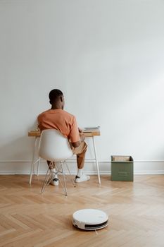 A man working at a desk in a minimalist home office with a robot vacuum cleaner on a wooden floor.
