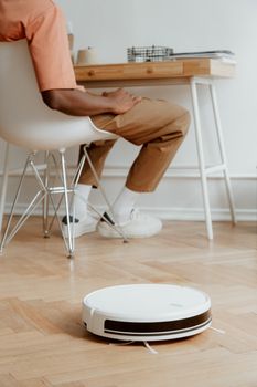 A man in casual attire sits as a robotic vacuum cleans the wooden floor.